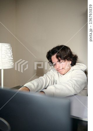 Teenage Boy Doing Homework At A Cozy Home Desk In The Evening Under Warm Lamp Light 136763839