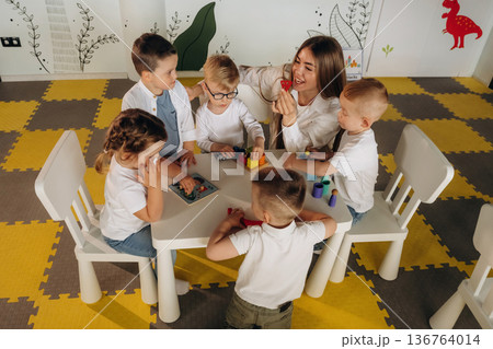 Sitting by the table. Children in kindergarten with their teacher Sitting by the table. Children in kindergarten with their teacher 136764014