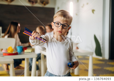 Boy in glasses is standing and holding pencils. Children in kindergarten with their teacher Boy in glasses is standing and holding pencils. Children in kindergarten with their teacher 136764093