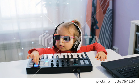 Young girl sits at home making music on a midi keyboard. She wears headphones and focuses on her creative process while enjoying her hobby Young girl sits at home making music on a midi keyboard. She wears headphones and focuses on her creative process while enjoying her hobby 136764134