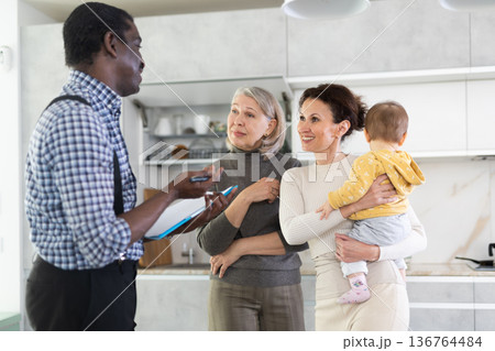 Two women with baby standing in the kitchen face to face with male agent Two women with baby standing in the kitchen face to face with male agent 136764484