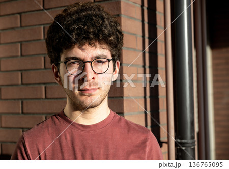 A young man with curly hair and glasses, contemplating a city view from a balcony, with buildings and distant mountains creating an urban background 136765095