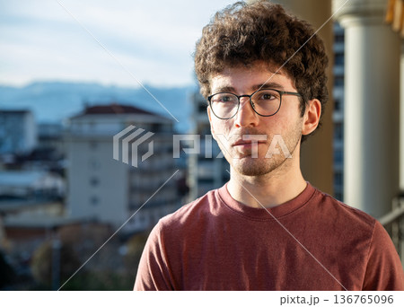 A young man with curly hair and glasses, contemplating a city view from a balcony, with buildings and distant mountains creating an urban background 136765096