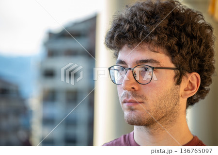 A young man with curly hair and glasses, contemplating a city view from a balcony, with buildings and distant mountains creating an urban background 136765097