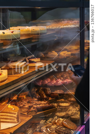 Bakery display case with cakes, cheesecake, pastry and chocolate dessert on shelves behind glass with customer holding smartphone. Concept of bakery shopping, dessert choice, food retail purchase Bakery display case with cakes, cheesecake, pastry and chocolate dessert on shelves behind glass with customer holding smartphone. Concept of bakery shopping, dessert choice, food retail purchase 136766017