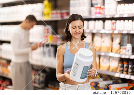 Positive woman choosing protein powder jar in sports nutrition store Positive woman choosing protein powder jar in sports nutrition store 136768859