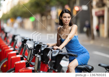 Brunette in blue dress rents electric bike on street of european city 136769101