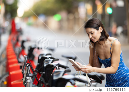 Brunette in blue dress rents electric bike - pays with mobile phone at street parking Brunette in blue dress rents electric bike - pays with mobile phone at street parking 136769113