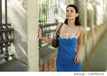 Cheerful brunette girl in blue dress walking along the street among architecture with bag Cheerful brunette girl in blue dress walking along the street among architecture with bag 136769132