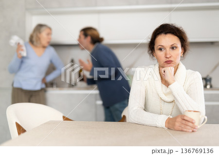 Sad thinking woman sits in kitchen and listens to man quarrel with elderly woman Sad thinking woman sits in kitchen and listens to man quarrel with elderly woman 136769136