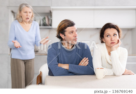 Elderly mother quarrels with adult thinking couple in bright kitchen Elderly mother quarrels with adult thinking couple in bright kitchen 136769140