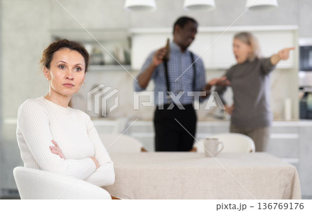 Middle-aged woman sitting at the kitchen table with her back to man and old woman quarreling together Middle-aged woman sitting at the kitchen table with her back to man and old woman quarreling together 136769176