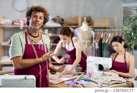 Middle-aged male dressmaker inviting clients while others sew and draw Middle-aged male dressmaker inviting clients while others sew and draw 136769177