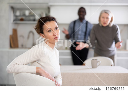 Middle-aged woman sitting at the kitchen table with her back to man and old woman quarreling to her Middle-aged woman sitting at the kitchen table with her back to man and old woman quarreling to her 136769233