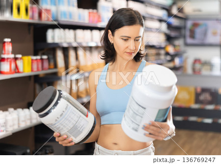 Focused woman choosing protein powder jar in sports nutrition store 136769247