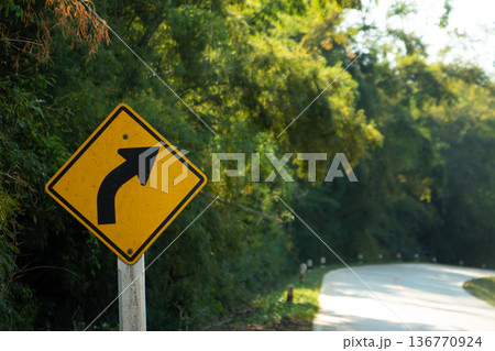 Right curve warning road sign beside a winding rural road surrounded by green trees in warm natural daylight. Right curve warning road sign beside a winding rural road surrounded by green trees in warm natural daylight. 136770924