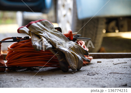 Work Gloves And Orange Extension Cord On Ground Near Vehicle In Outdoor Workshop Work Gloves And Orange Extension Cord On Ground Near Vehicle In Outdoor Workshop 136774231