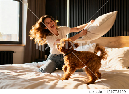 Young caucasian female enjoying pillow fight with dog on bed in sunlit room 136778469