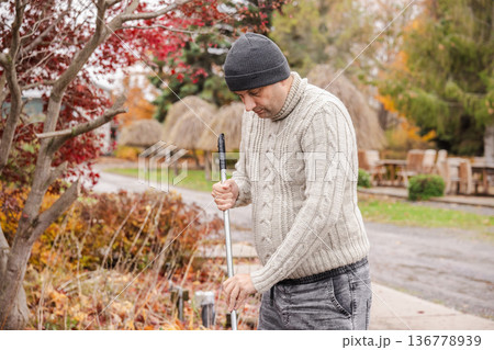 Male small business owner raking fallen autumn leaves near outdoor cafe terrace while preparing seasonal restaurant area for closing 136778939