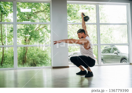 Lifts a kettlebell. Strong man is indoors in the gym with panoramic windows 136779390