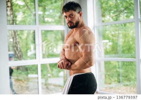 Shows the muscles. Shirtless strong man is standing against panoramic windows Shows the muscles. Shirtless strong man is standing against panoramic windows 136779397
