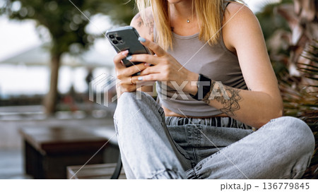 Woman sits outside while using smartphone in a casual setting during daytime Woman sits outside while using smartphone in a casual setting during daytime 136779845