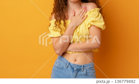 Person in yellow top stands against a bright orange background with hands on chest Person in yellow top stands against a bright orange background with hands on chest 136779882