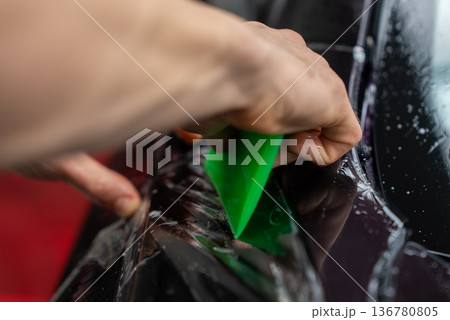 A detailer's hand uses a bright green squeegee to smooth clear paint protection film on a glossy black panel. Beaded water shows wet application in a premium auto studio. 136780805