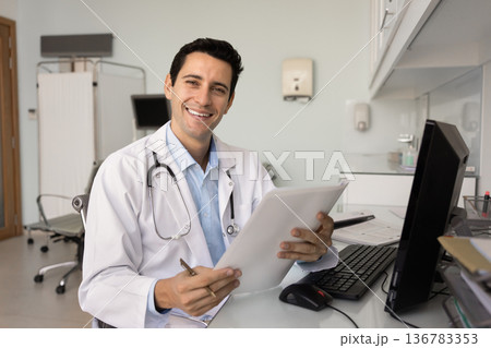 Portrait of doctor looks at camera, posing seated at desk Portrait of doctor looks at camera, posing seated at desk 136783353