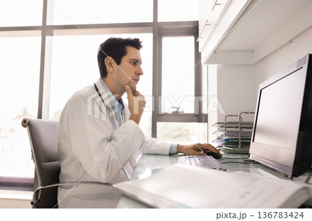 Young 30s male doctor sits at desk working on computer Young 30s male doctor sits at desk working on computer 136783424