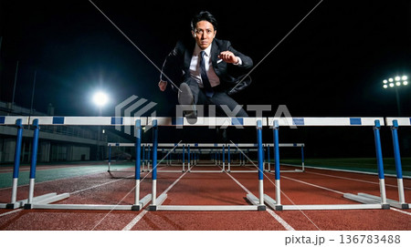 夜の競技場で障害を乗り越え飛躍するスーツ姿の男性のビジネスイメージ 136783488
