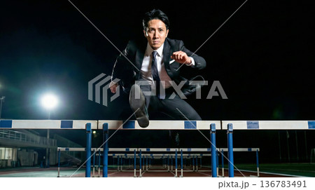 夜の競技場で障害を乗り越え飛躍するスーツ姿の男性のビジネスイメージ 136783491