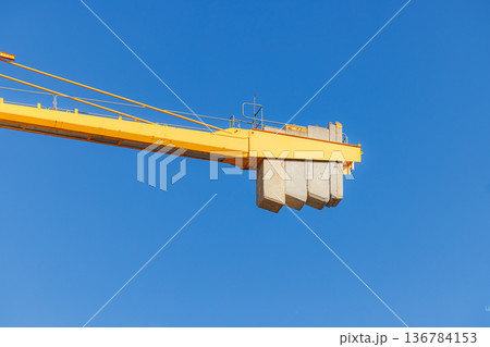 Large yellow crane lifting concrete blocks against a vibrant blue sky at a construction site Large yellow crane lifting concrete blocks against a vibrant blue sky at a construction site 136784153
