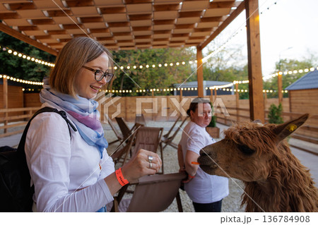 Woman feeding a friendly llama in a cozy outdoor space during golden hour Woman feeding a friendly llama in a cozy outdoor space during golden hour 136784908