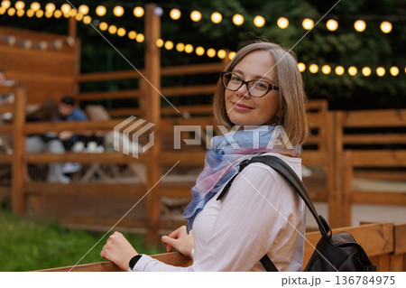Smiling woman enjoying a peaceful evening surrounded by warm lights in a cozy outdoor setting 136784975