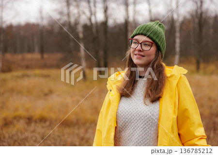 Smiling woman in a yellow raincoat enjoys a crisp autumn day in a tranquil park setting 136785212
