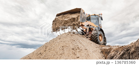 A powerful bulldozer or loader moves large volumes of sand at a busy construction site under a dramatic sky covered with gray clouds. Excavation work A powerful bulldozer or loader moves large volumes of sand at a busy construction site under a dramatic sky covered with gray clouds. Excavation work 136785360