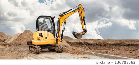 A mini excavator digs the earth, shaping the landscape at a busy construction site. Using compact equipment for construction work A mini excavator digs the earth, shaping the landscape at a busy construction site. Using compact equipment for construction work 136785362