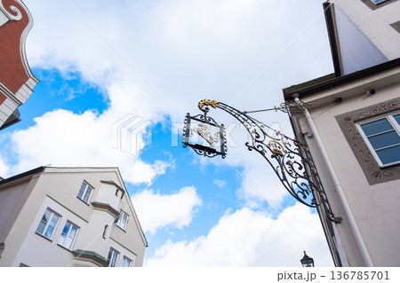 A decorative metal bracket hangs above colorful houses in Memmingen, Germany. The scene features vibrant architecture under a blue sky with clouds. A decorative metal bracket hangs above colorful houses in Memmingen, Germany. The scene features vibrant architecture under a blue sky with clouds. 136785701
