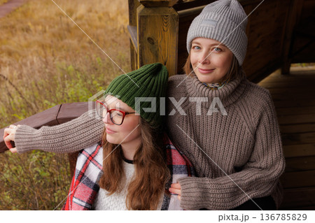 Friends enjoy cozy autumn vibes together while wrapped in warm sweaters on a wooden porch in nature Friends enjoy cozy autumn vibes together while wrapped in warm sweaters on a wooden porch in nature 136785829