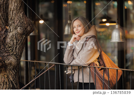 Charming woman in stylish coat enjoying a quiet moment outside a cozy cafe in the early morning light Charming woman in stylish coat enjoying a quiet moment outside a cozy cafe in the early morning light 136785839