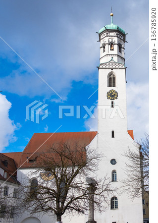 The Hospital Church of St. Peter and Paul in Memmingen, Germany, features a tall clock tower, surrounded by trees and traditional buildings under a blue sky with clouds. 136785920