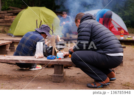Campers gather around the fire, preparing a warm meal in a serene outdoor setting Campers gather around the fire, preparing a warm meal in a serene outdoor setting 136785948