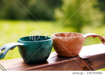 Beautiful ceramic cups resting on a wooden railing under the warm sunlight in a tranquil outdoor setting 136785960