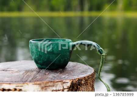 Green ceramic cup resting on a wooden log beside a peaceful lake during a rainy afternoon 136785999