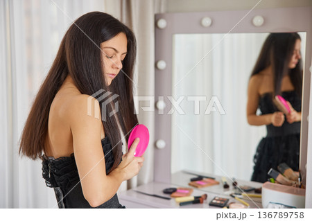 Woman standing in front of illuminated mirror, brushing her long, dark hair Woman standing in front of illuminated mirror, brushing her long, dark hair 136789758