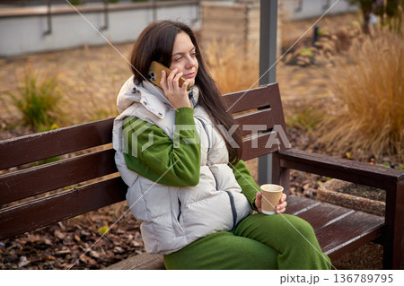 Woman having an outdoor phone conversation while sitting on a bench with coffee Woman having an outdoor phone conversation while sitting on a bench with coffee 136789795