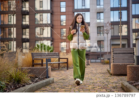 stylish woman walking outdoors, holding a coffee cup and looking at her smartphone stylish woman walking outdoors, holding a coffee cup and looking at her smartphone 136789797