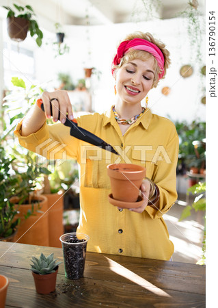 A smiling woman carefully tends to a plant, adding soil to a small pot with a trowel A smiling woman carefully tends to a plant, adding soil to a small pot with a trowel 136790154