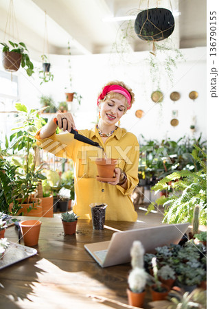 A woman in a yellow shirt and pink headscarf is planting succulents in a terracotta pot, surrounded by lush greenery 136790155